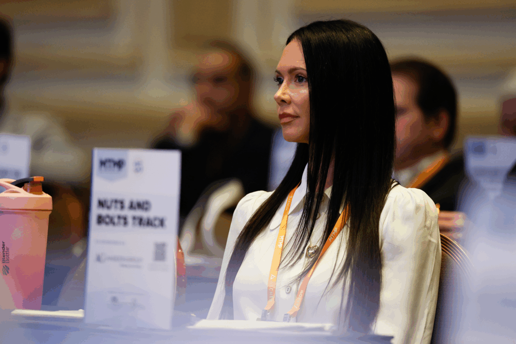 A woman wearing a conference badge sits in a meeting room, listening attentively while facing forward.