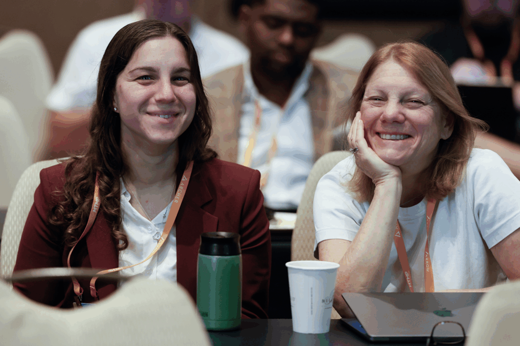 Two women wearing conference badges sit side by side at a table, smiling and listening during a conference session.