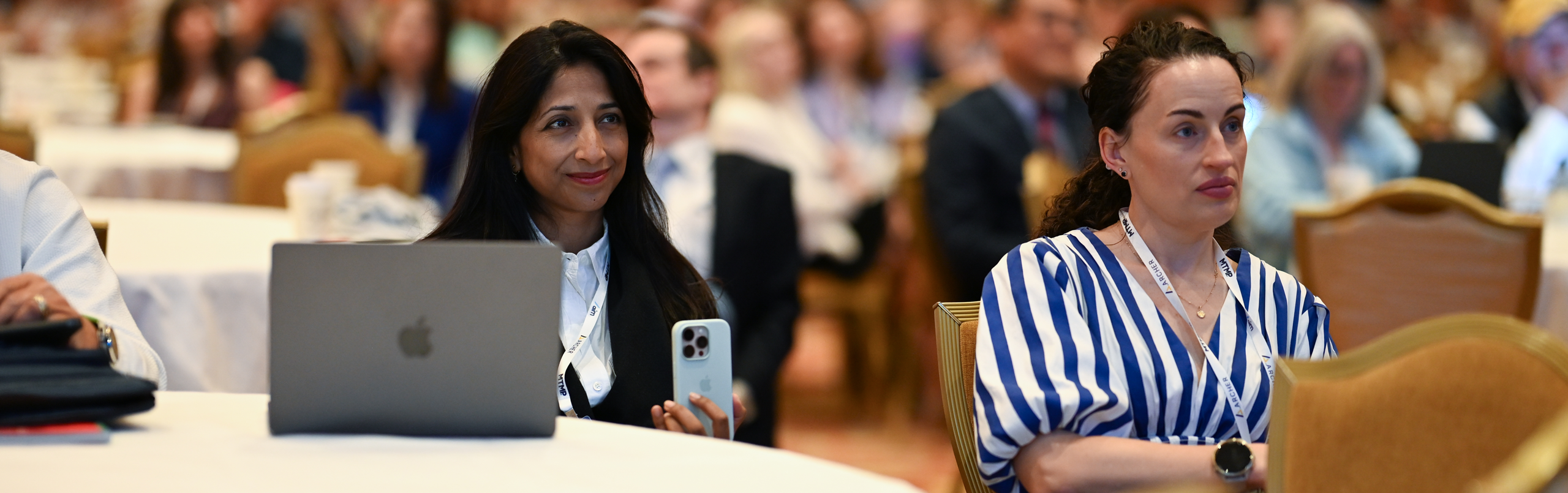 Two women sit at a round table facing forward during a conference, with one woman looking at a laptop and holding a smartphone.